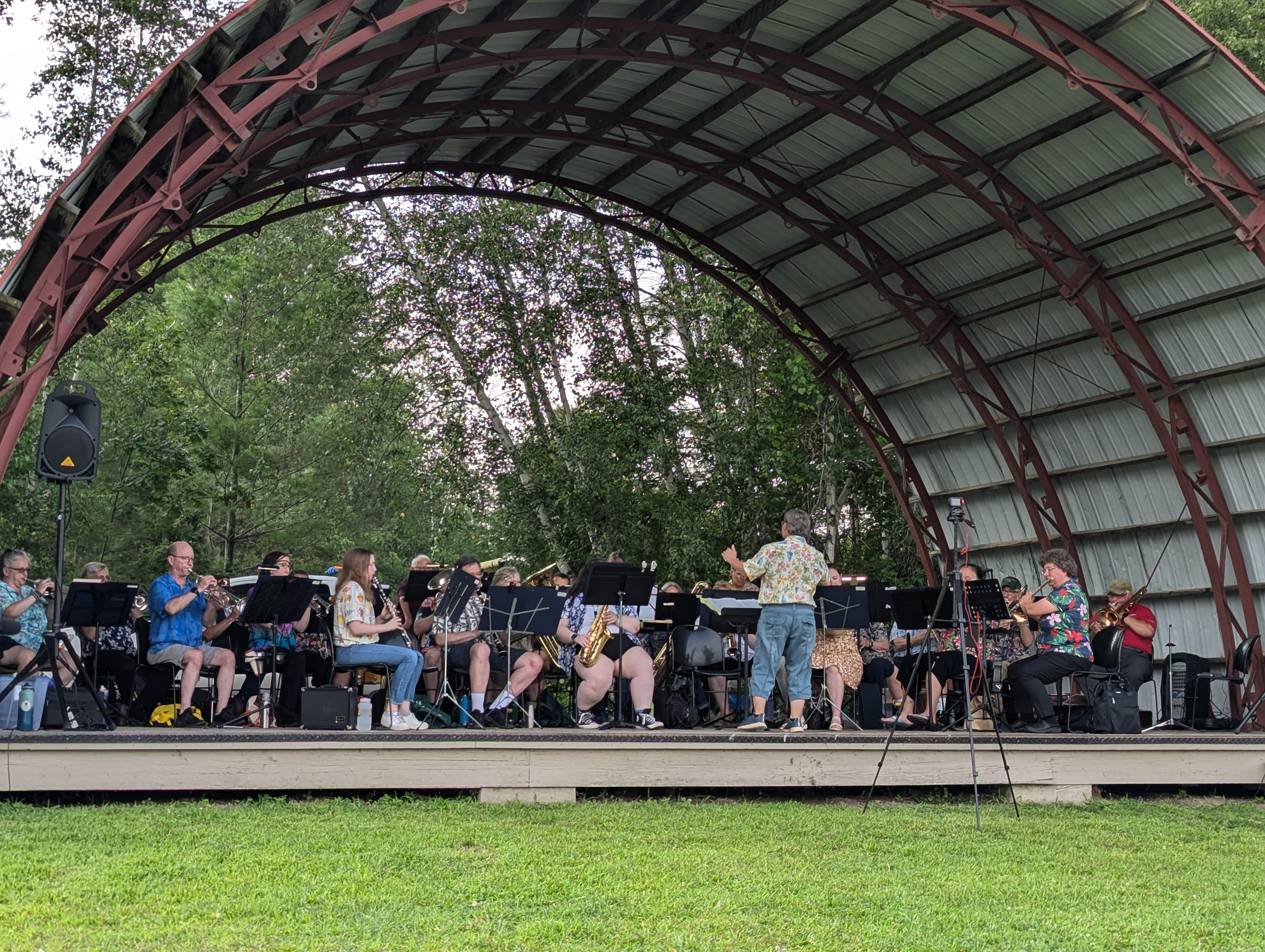 Picture of the band performing outdoors under a performance shell with trees behind them